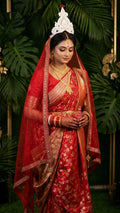 A bengali bride wearing Red Pure Katan Silk saree with floral meenakari work in a garden.