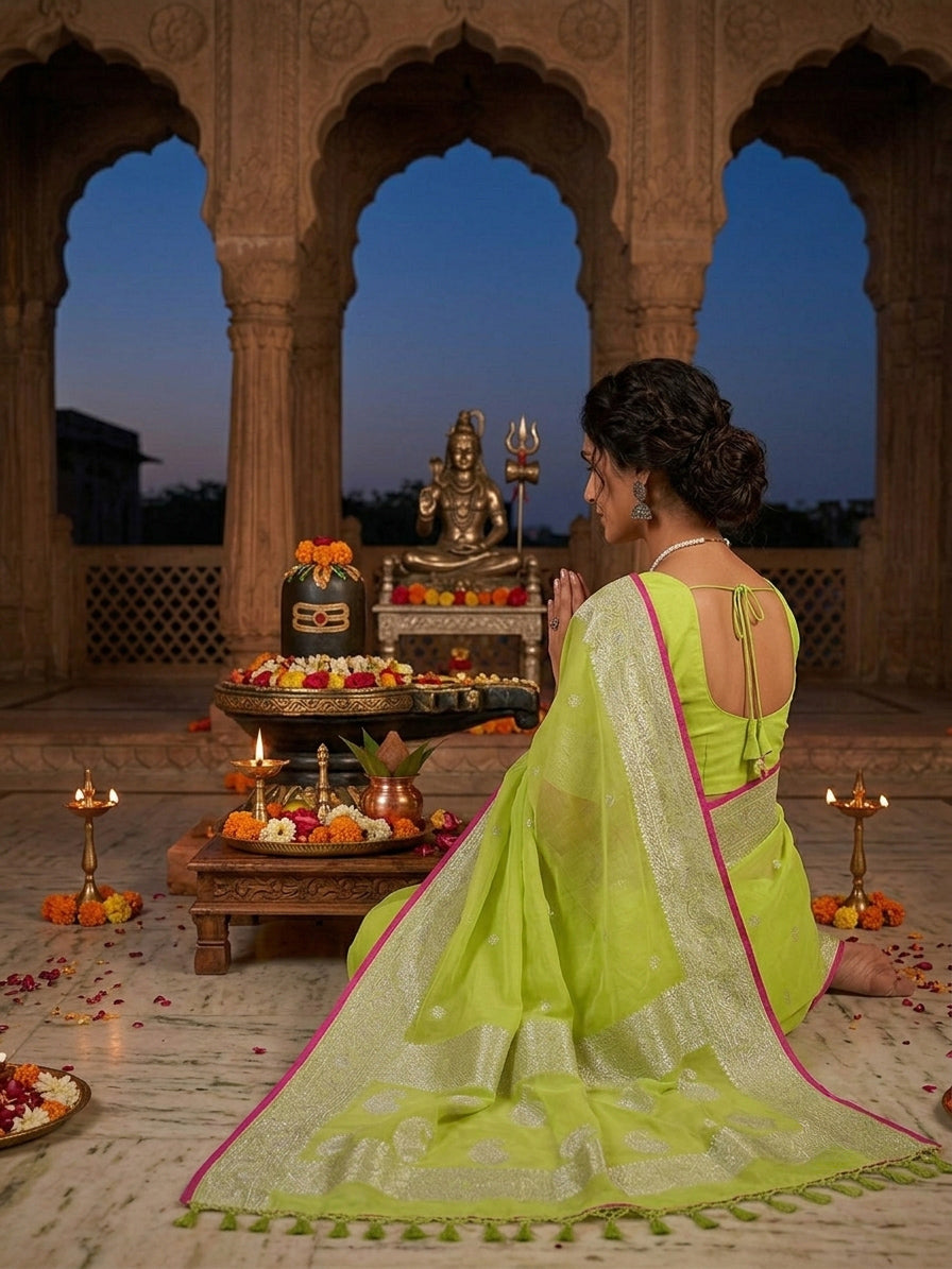 A woman in a bright lime green saree with a silver zari border and pallu, kneeling in front of a temple altar with a brass idol. The background features stone arches and a twilight sky.